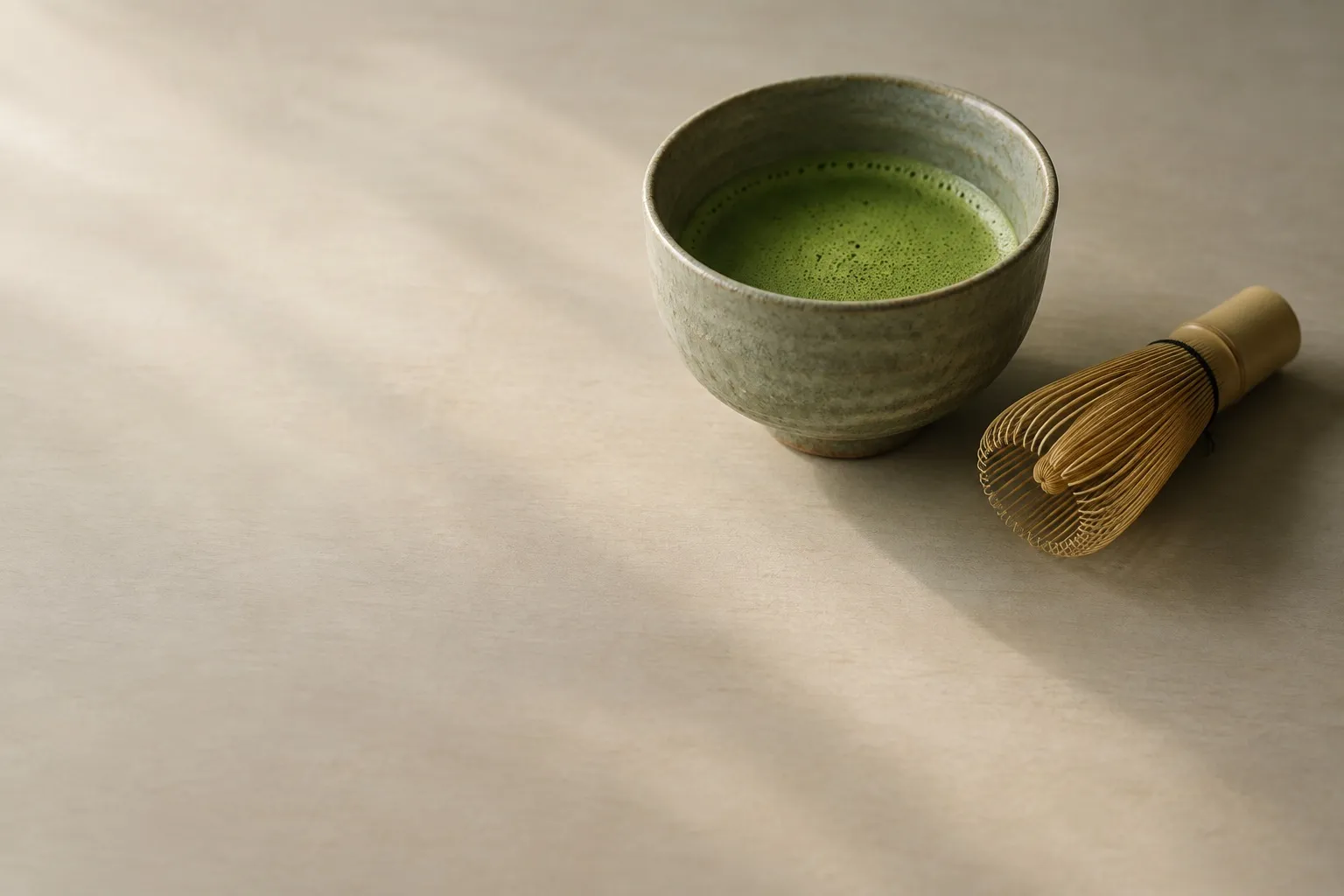 A matcha bowl and a bamboo whisk on a bare table, in low daylight.